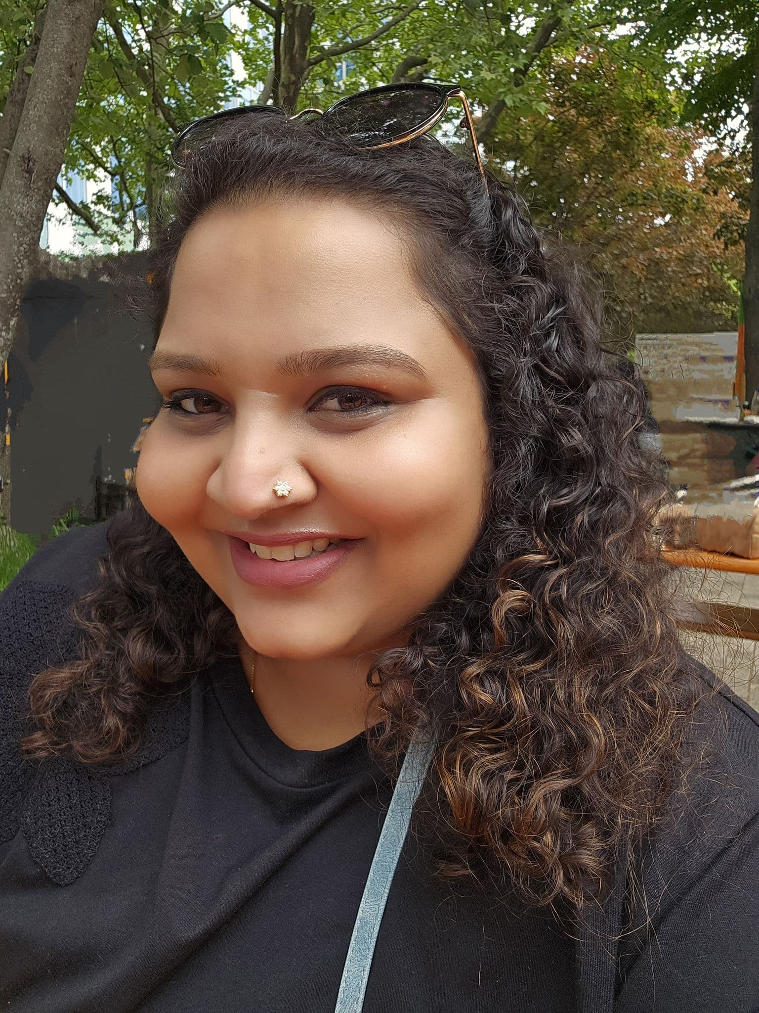Smiling woman with dark curly hair wearing a nose stud and sunglasses on her head.